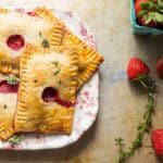 Presentation shot showing the finished pastries on a plate with a decorative floral rim. Fresh strawberries are arranged around the plate and in a teal container visible at the edge of the frame.