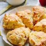 Golden-brown goat cheese and prosciutto scones arranged on a white scalloped plate. A small bowl of pink strawberry butter is visible in the background along with fresh strawberries on a marble countertop.