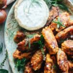 A close-up of crispy tomato fritters stacked generously around a dish of herbed tzatziki sauce. Flecks of sea salt and sprigs of dill and mint highlight the dish's vibrant, savory charm.