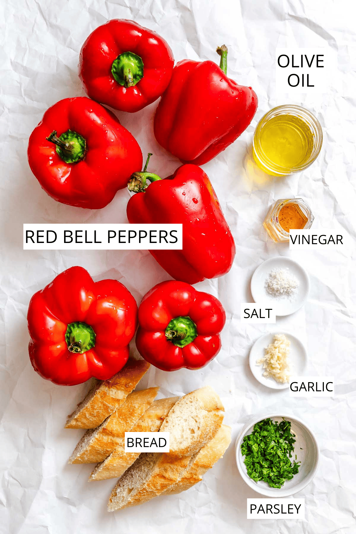 Flat lay of ingredients for roasted red peppers, including whole red bell peppers, sliced bread, garlic, salt, parsley, olive oil, and vinegar labeled for clarity.
