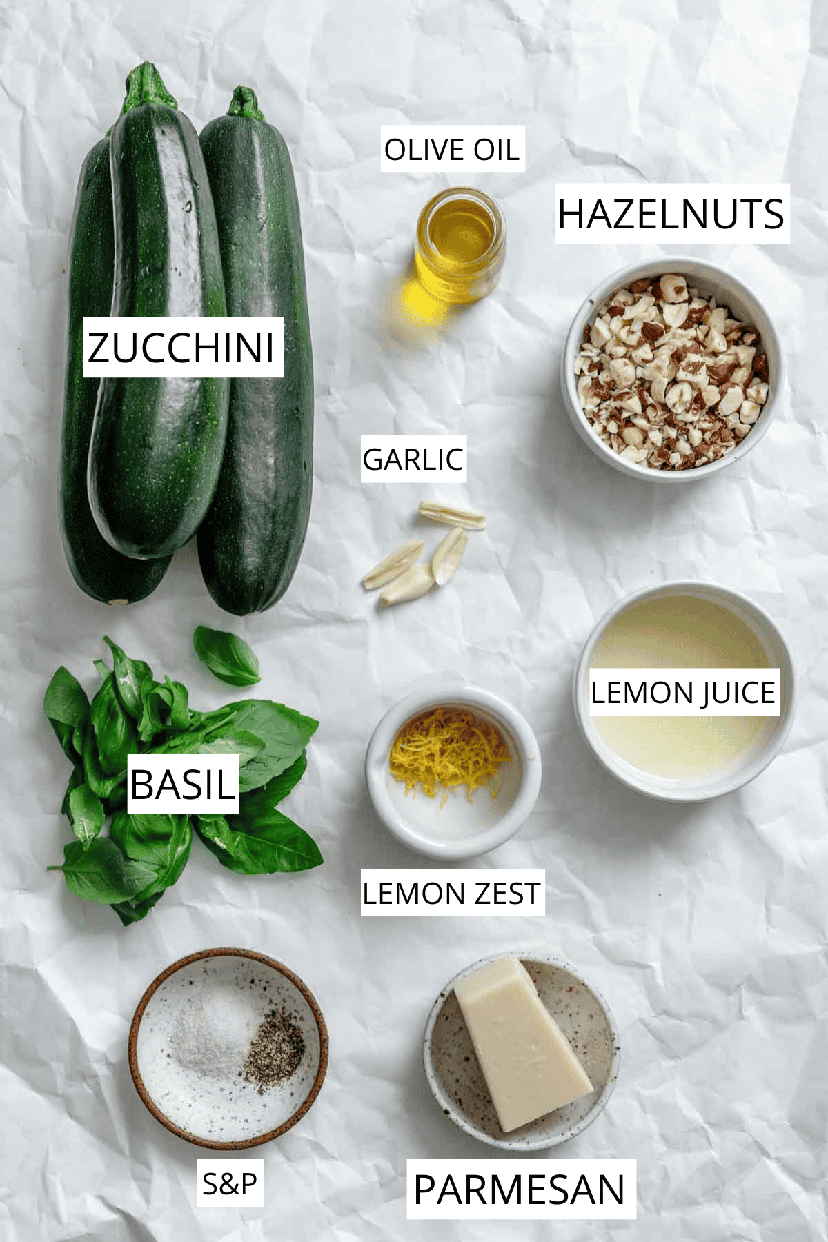 Overhead shot of recipe ingredients on white parchment paper, each labeled - fresh zucchini, basil leaves, chopped hazelnuts, parmesan cheese, lemon juice, olive oil, garlic cloves, lemon zest, and salt and pepper in small bowls and dishes.