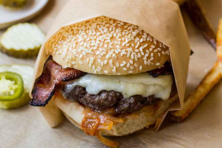Close-up of a bacon cheddar burger wrapped in parchment, featuring a sesame seed bun, crispy bacon, melted cheese, and caramelized tomato onion jam.