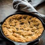 Freshly baked skillet chocolate chip cookie before adding toppings, displaying its perfectly golden surface studded with chocolate chunks and a light sprinkle of sea salt. A gray linen napkin frames the cast iron pan on a dark wooden surface.
