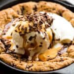 Close-up of a freshly baked chocolate chip cookie in a cast iron skillet topped with vanilla ice cream, chocolate shavings, and caramel sauce. The warm cookie edges are golden-brown and crisp while the center remains soft beneath the melting ice cream.