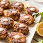 Angled view of fish cakes arranged in rows on a rectangular platter, showing their uniform size and consistent browning. Fresh herbs and lemon wedges adorn the plate, with a small dish of dipping sauce visible in the background.
