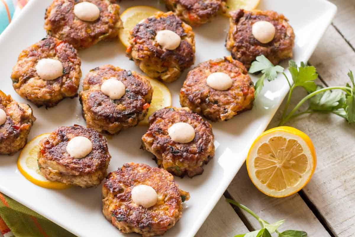 Golden-brown mini fish cakes arranged on a rectangular white platter, each topped with a creamy aioli dollop. Thin lemon slices and fresh parsley sprigs garnish the plate, emphasizing their appetizer presentation.