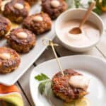 Elegant serving setup with a platter of fish cakes alongside individual portions and dipping sauce. One fish cake speared with a wooden pick sits on a small plate with a lemon slice, while a small bowl of creamy sauce with wooden spoon accompanies the arrangement.