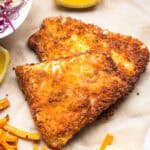 An overhead view of golden-brown, triangular panko-crusted fish fillets on parchment paper. The crispy coating has a rich amber color with darker spots where it's most deeply fried. Lemon wedges are placed around the fish, with sweet potato fries visible at the bottom of the frame and a small portion of coleslaw with purple cabbage visible in the upper left corner.