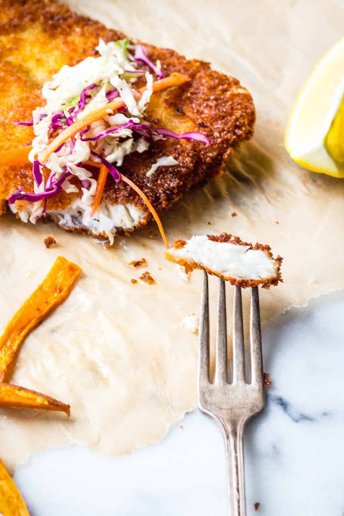 A close-up image showing a bite being taken from the panko-crusted fish, with a fork holding a small piece. The white, flaky interior of the fish is visible in contrast to the crispy golden exterior. Some coleslaw has been placed on top of one piece of fish, and a lemon wedge sits nearby. This image demonstrates the texture contrast between the crunchy coating and tender fish inside.