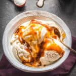 Overhead view of a bowl of vanilla ice cream topped with a thick drizzle of homemade salted caramel sauce, set on a dark surface with scattered sea salt and a jar of caramel in the background.