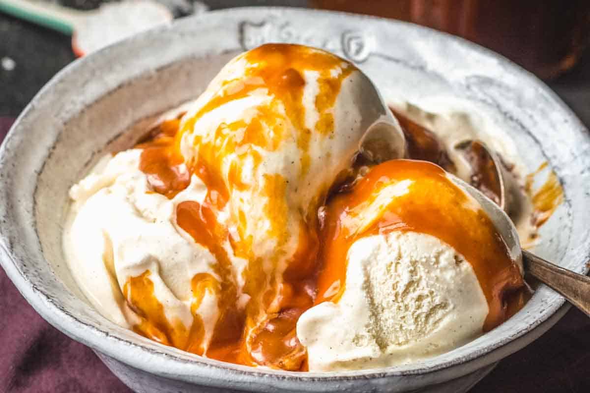 Close-up of scoops of vanilla ice cream in a rustic ceramic bowl, generously drizzled with a thick, golden homemade salted caramel sauce.