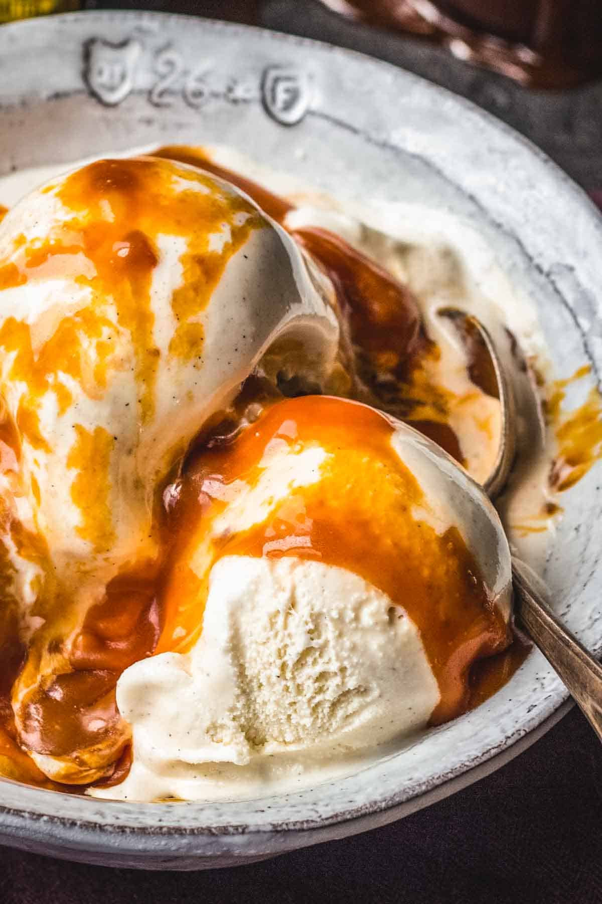 A bowl of vanilla ice cream with visible vanilla bean specks, topped with rich, glossy ribbons of salted caramel sauce and served with a tarnished silver spoon.