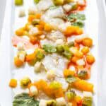 A delicate arrangement of crudo topped with colorful diced fruit and vegetables, including golden mango, green kiwi, and white jicama, with splashes of red chili oil and fresh cilantro on a white serving dish.