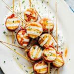 Close-up view of grilled prosciutto-wrapped scallops on wooden skewers. The caramelized scallops with crispy prosciutto are arranged on a white oval plate with a rustic rim, sitting in a pool of creamy sauce with olive oil drizzled on top.