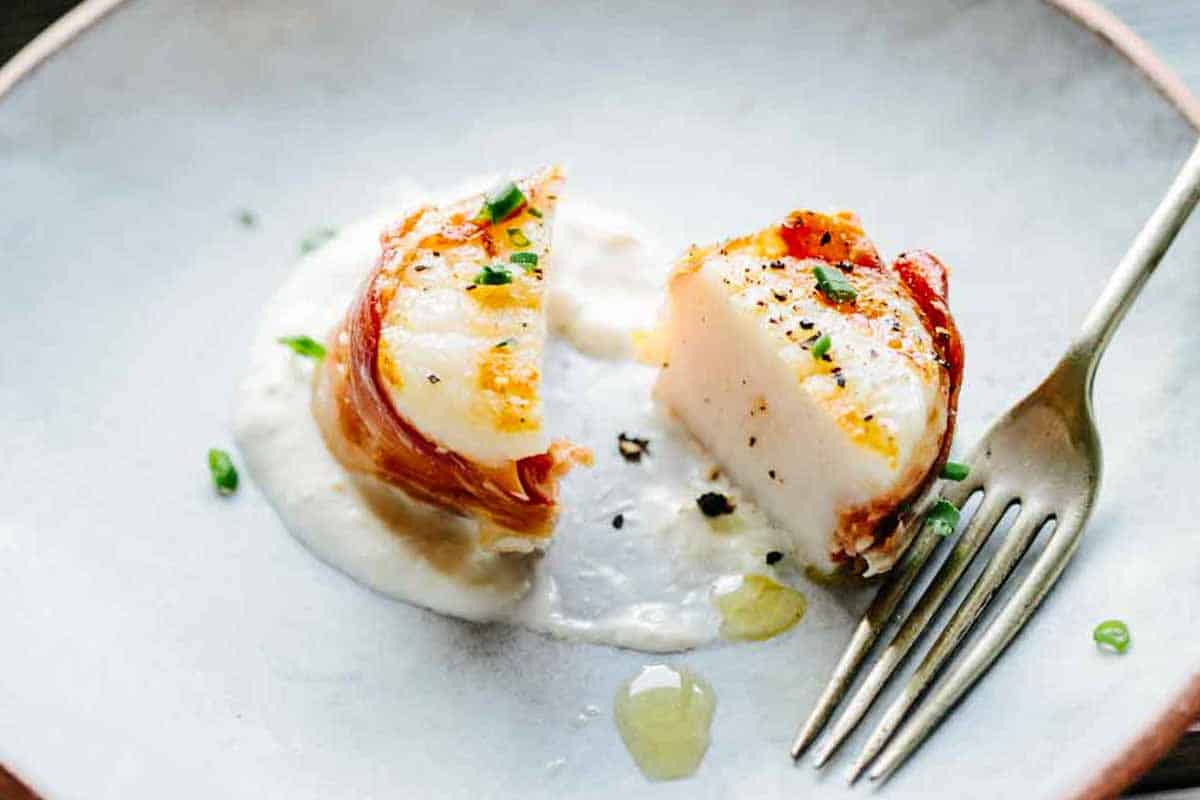 A close-up of a single prosciutto-wrapped scallop that has been cut in half, showing the tender interior of the scallop, the thin layer of crispy prosciutto around it, and the creamy sauce underneath, with a fork in the frame.
