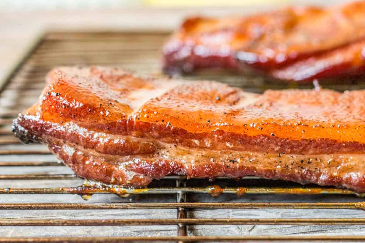 Close-up of thick-cut bacon displaying distinct layers of fat and meat. The fat has turned translucent while the meat portion shows a rich pink-red color with black pepper flecks visible on the surface.