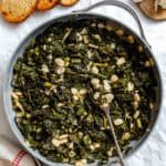 Finished Tuscan kale and beans dish served in a large skillet, with toasted bread slices and garlic cloves on a wooden board alongside. A metal spoon rests in the skillet, showing the moist, saucy consistency of the greens and beans.
