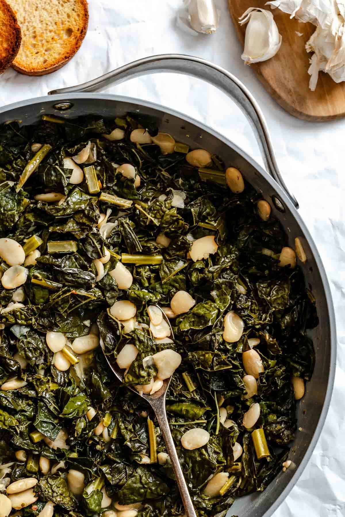 Close-up overhead view of the Tuscan kale and white beans in the skillet, showing more detail of the tender greens, plump beans, and glistening olive oil. Toasted bread and garlic are visible at the edge of the frame.
