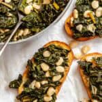 Tuscan kale and bean mixture being served on toasted bread slices, creating crostini. The serving skillet appears in the upper left corner while several topped bread slices are arranged on parchment paper.