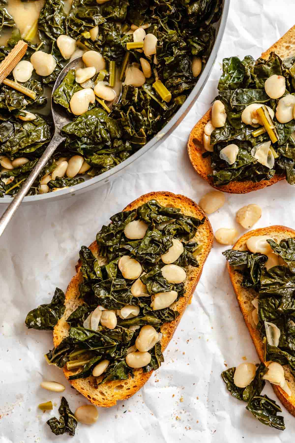 Tuscan kale and bean mixture being served on toasted bread slices, creating crostini. The serving skillet appears in the upper left corner while several topped bread slices are arranged on parchment paper.