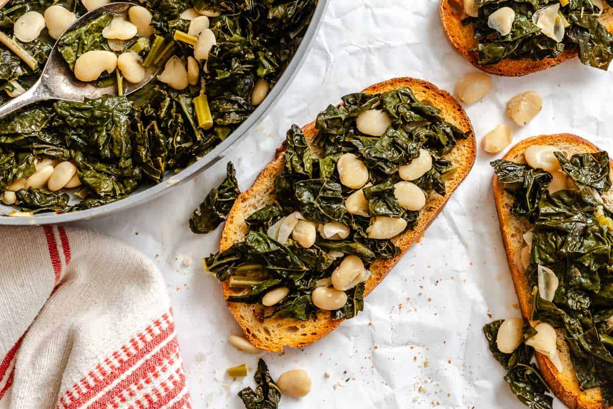 Similar angle to Image 6, showing the skillet of kale and beans with a serving spoon alongside several pieces of topped crostini on parchment paper. A red-striped kitchen towel is visible in the corner.