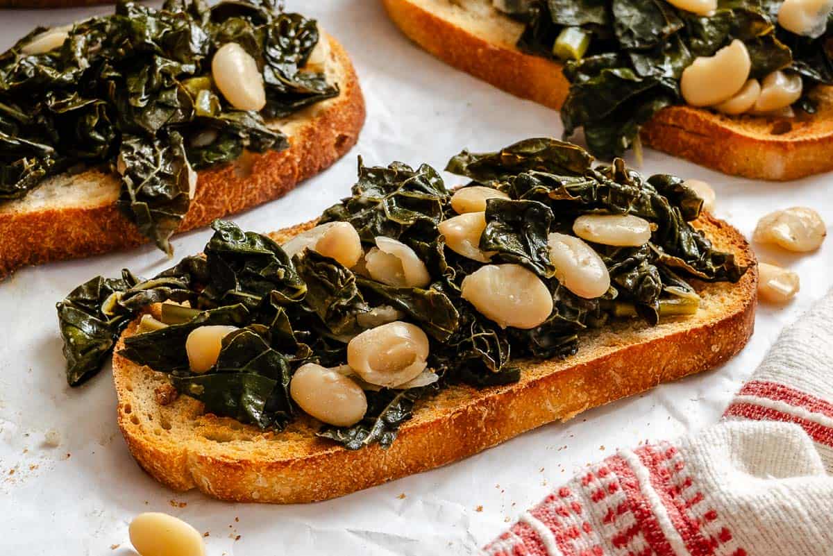 Close-up of several pieces of toasted bread topped with the Tuscan kale and white bean mixture. The image highlights the textural contrast between the crisp bread and the tender greens and beans, with some beans scattered on the paper.