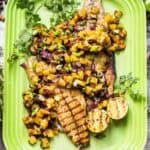 Grilled fish fillet on a bright lime-green Le Creuset platter, topped with vibrant yellow pineapple salsa featuring red onions, green scallions, and fresh cilantro. Charred lime halves accompany the dish, which is presented on newspaper.