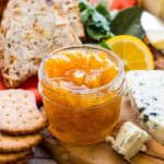 Meyer lemon marmalade in a glass container surrounded by crackers, brown bread, and different kinds of cheeses.