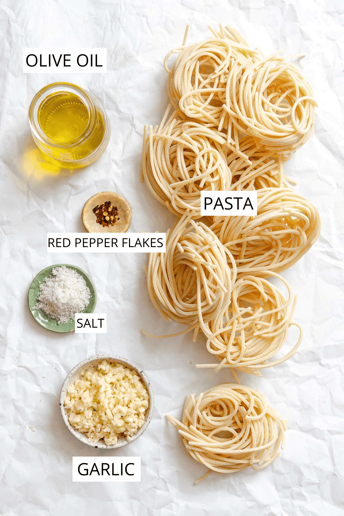 Ingredients for pasta aglio e olio arranged on white parchment paper, featuring nests of uncooked spaghetti on the right, alongside small dishes containing olive oil, red pepper flakes, finely minced garlic.