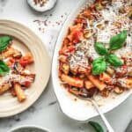 A table setting with two portions of pasta alla norma - one in a small beige plate and another in the white oval serving dish. Both are garnished with basil and cheese, creating an inviting presentation for sharing.