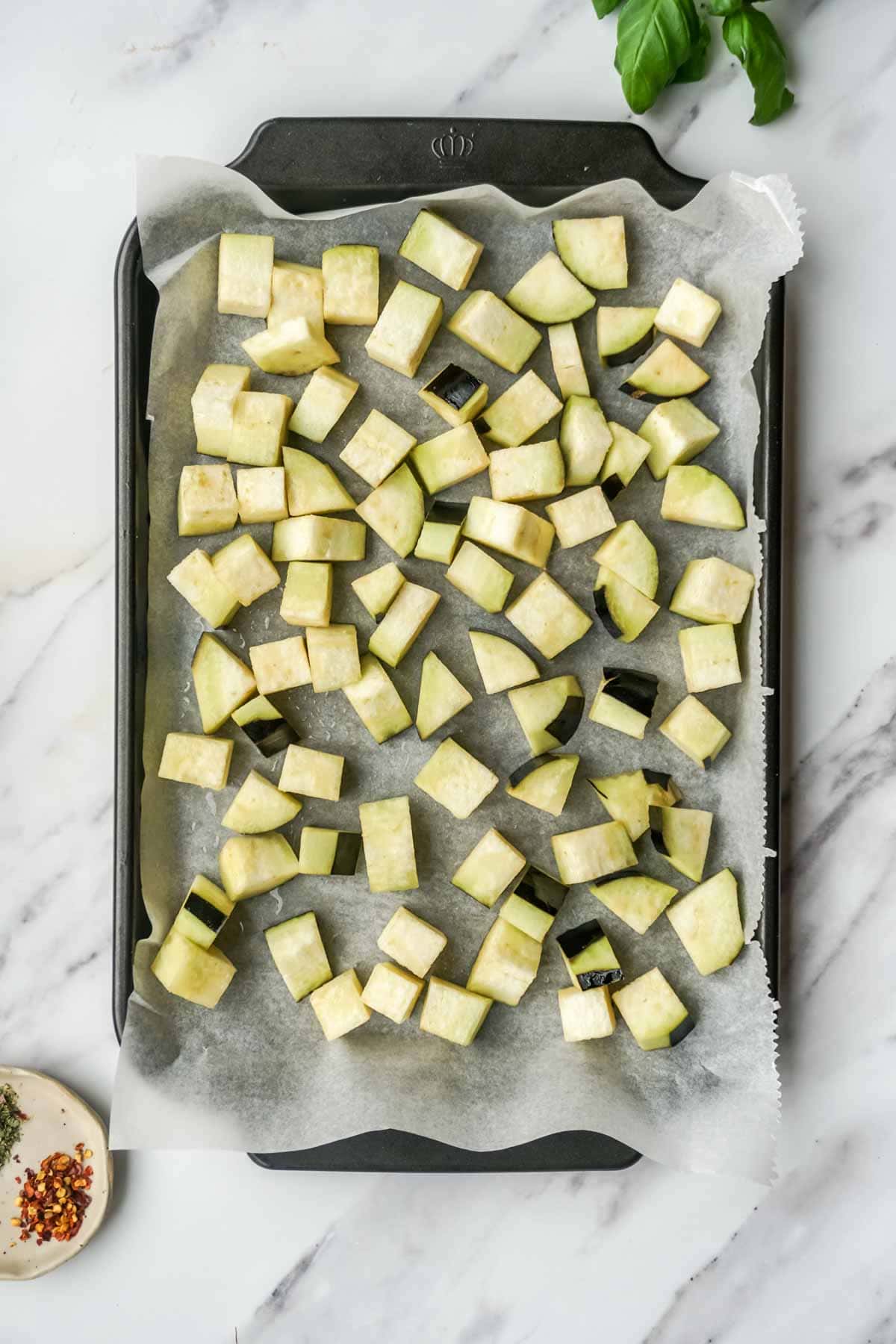 Raw diced eggplant arranged on a parchment-lined baking sheet. The uniformly cut cubes are spread evenly across the surface, ready for roasting. Fresh basil leaves and a small dish of red pepper flakes are visible in the corner, set against a white marble countertop.