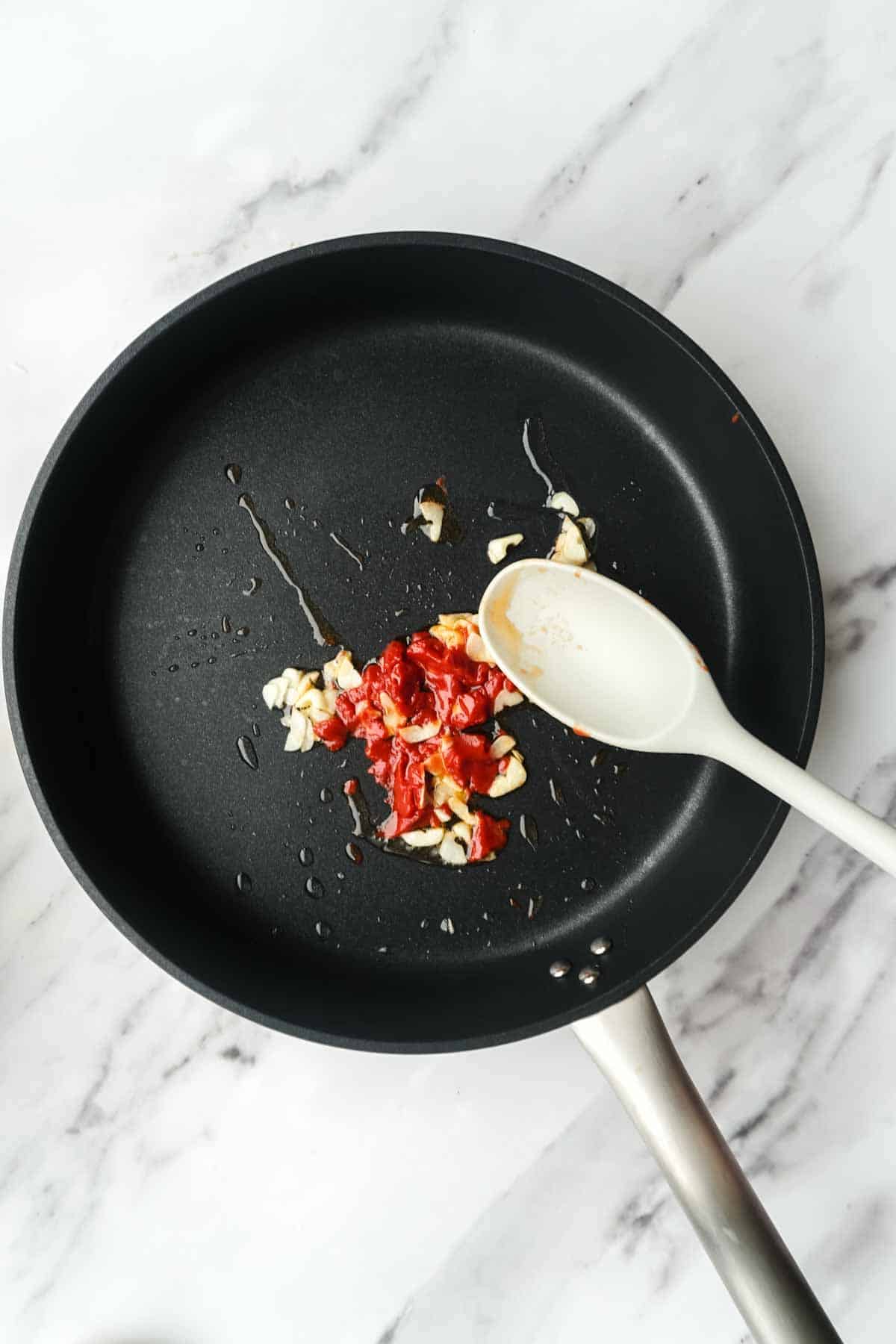 A black non-stick pan containing minced garlic and what appears to be tomato paste being sautéed. A white wooden spoon rests in the pan, showing the beginning stages of the sauce preparation on a marble surface.