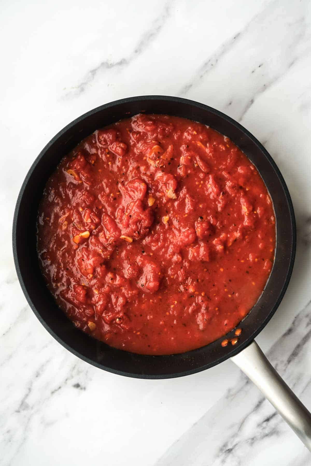Overhead view of a rich tomato sauce simmering in a black pan. The sauce has a vibrant red color and shows chunks of crushed tomatoes with visible seasonings, forming the base for the pasta alla norma.