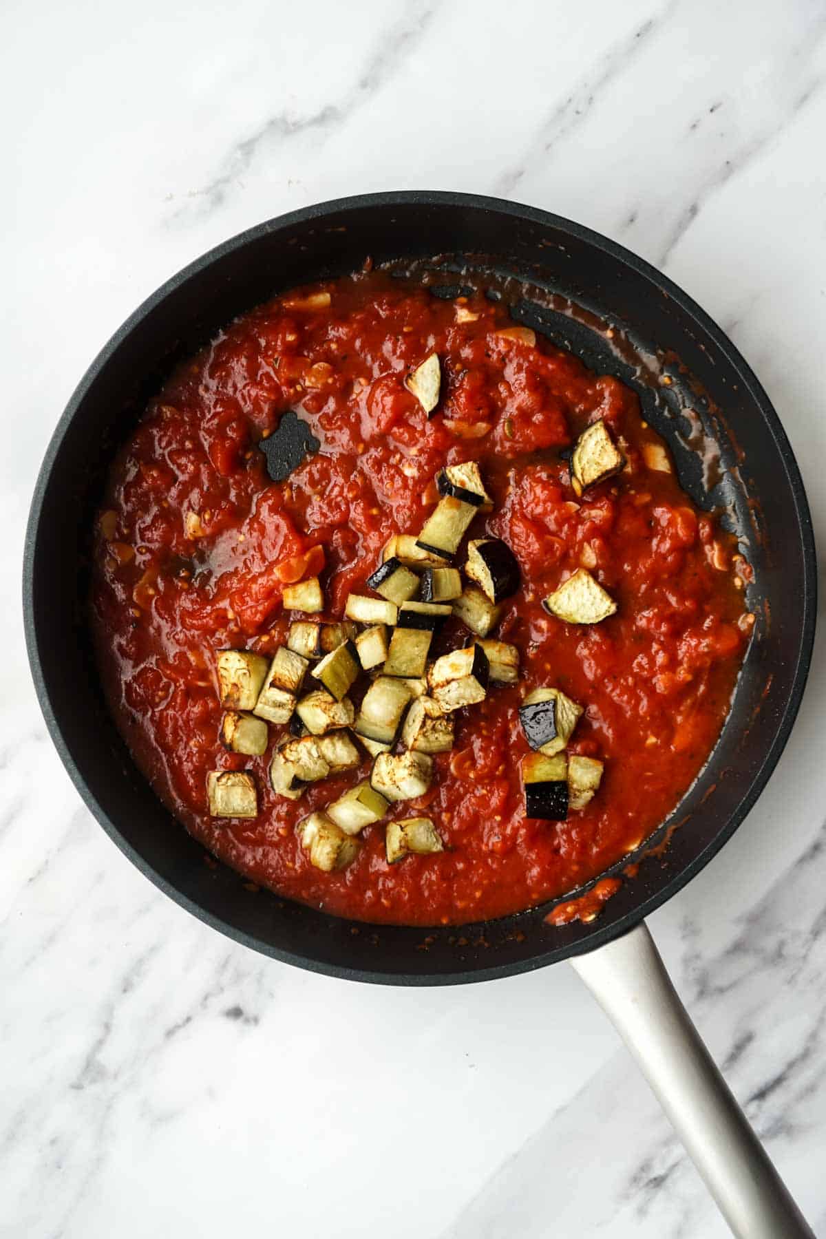 The tomato sauce in a black pan with roasted eggplant cubes being added. This captures the moment the two main components of the dish are combined, with the browned eggplant pieces sitting on top of the rich red sauce.