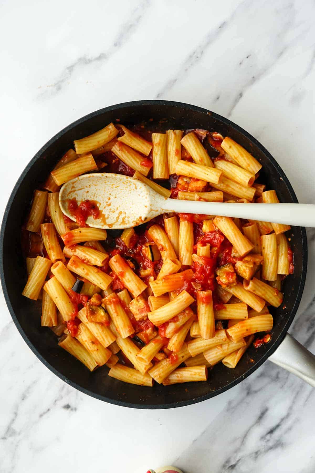 A black pan containing rigatoni pasta tossed with tomato sauce and roasted eggplant. A white wooden spoon shows the pasta being mixed with the sauce, creating the classic pasta alla norma before serving.