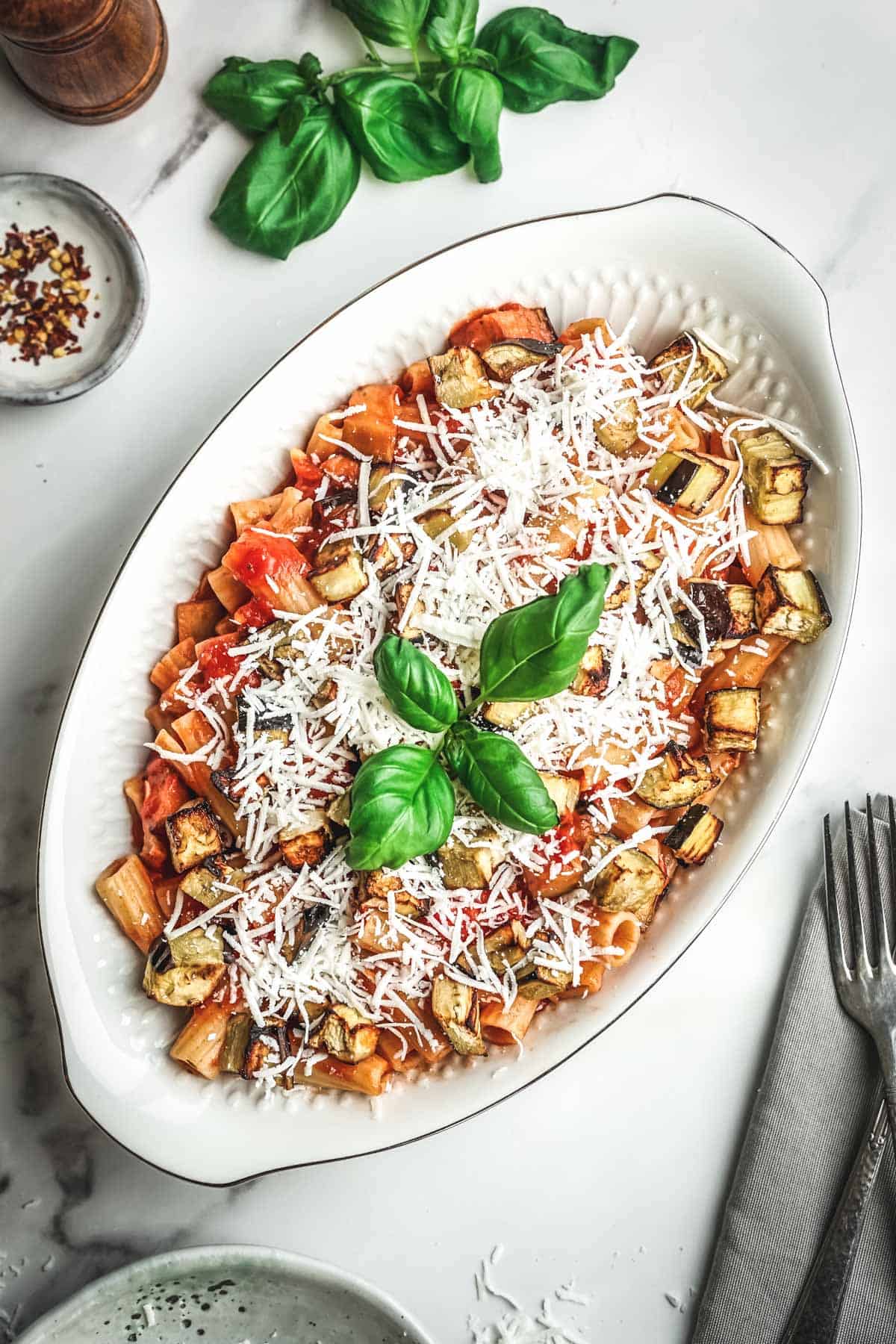 The finished pasta alla norma served in a white oval dish, garnished with shredded ricotta salata cheese and fresh basil leaves. The combination of tube pasta, eggplant pieces, and tomato sauce is complemented by the white cheese, creating an appetizing presentation.
