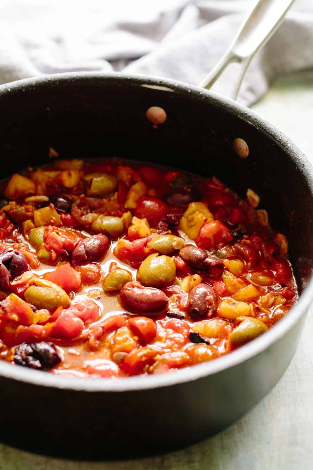 A saucepan containing a vibrant mixture of beans and tomatoes simmering in a rich sauce. Various types of beans are visible - including kidney beans, green olives, and chickpeas - swimming in a chunky tomato base that will serve as the sauce for the pasta dish.