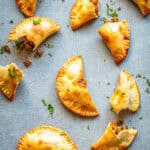 Golden-brown baked empanadas arranged on a light blue textured surface. The half-moon pastries have crimped edges and are garnished with chopped fresh herbs. Some are broken open, revealing the savory picadillo filling of seasoned ground beef with visible pieces of olives.