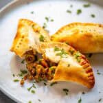 Three empanadas arranged on a white plate with one broken open to showcase the picadillo filling. The ground beef mixture contains visible pieces of olives and is garnished with fresh herbs. The empanada's flaky crust provides a perfect contrast to the savory filling.