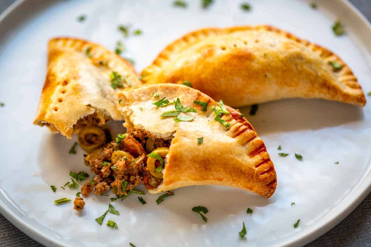 Similar to Image 4, showing three empanadas on a white plate with one broken open, displaying the picadillo filling with its distinctive mixture of ground beef, olives, and seasonings. Fresh herbs garnish both the plate and the tops of the empanadas.