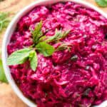 Vibrant magenta beet salad in a white bowl garnished with fresh mint leaves and dill fronds. The creamy texture suggests the addition of tahini, creating a rich contrast against the earthy sweetness of the shredded beets. The bowl sits on a wooden cutting board with scattered herbs.