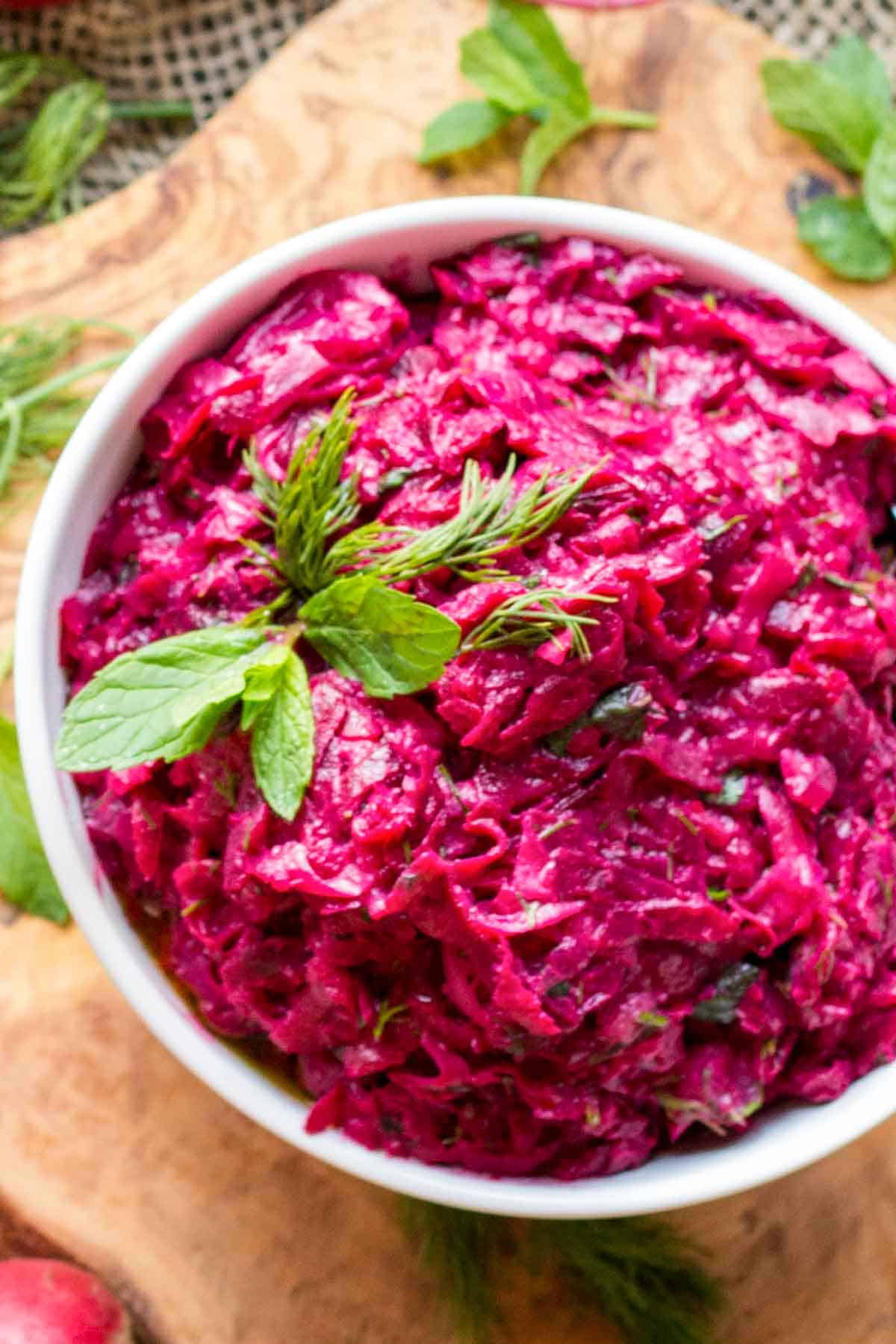 Vibrant magenta beet salad in a white bowl garnished with fresh mint leaves and dill fronds. The creamy texture suggests the addition of tahini, creating a rich contrast against the earthy sweetness of the shredded beets. The bowl sits on a wooden cutting board with scattered herbs.