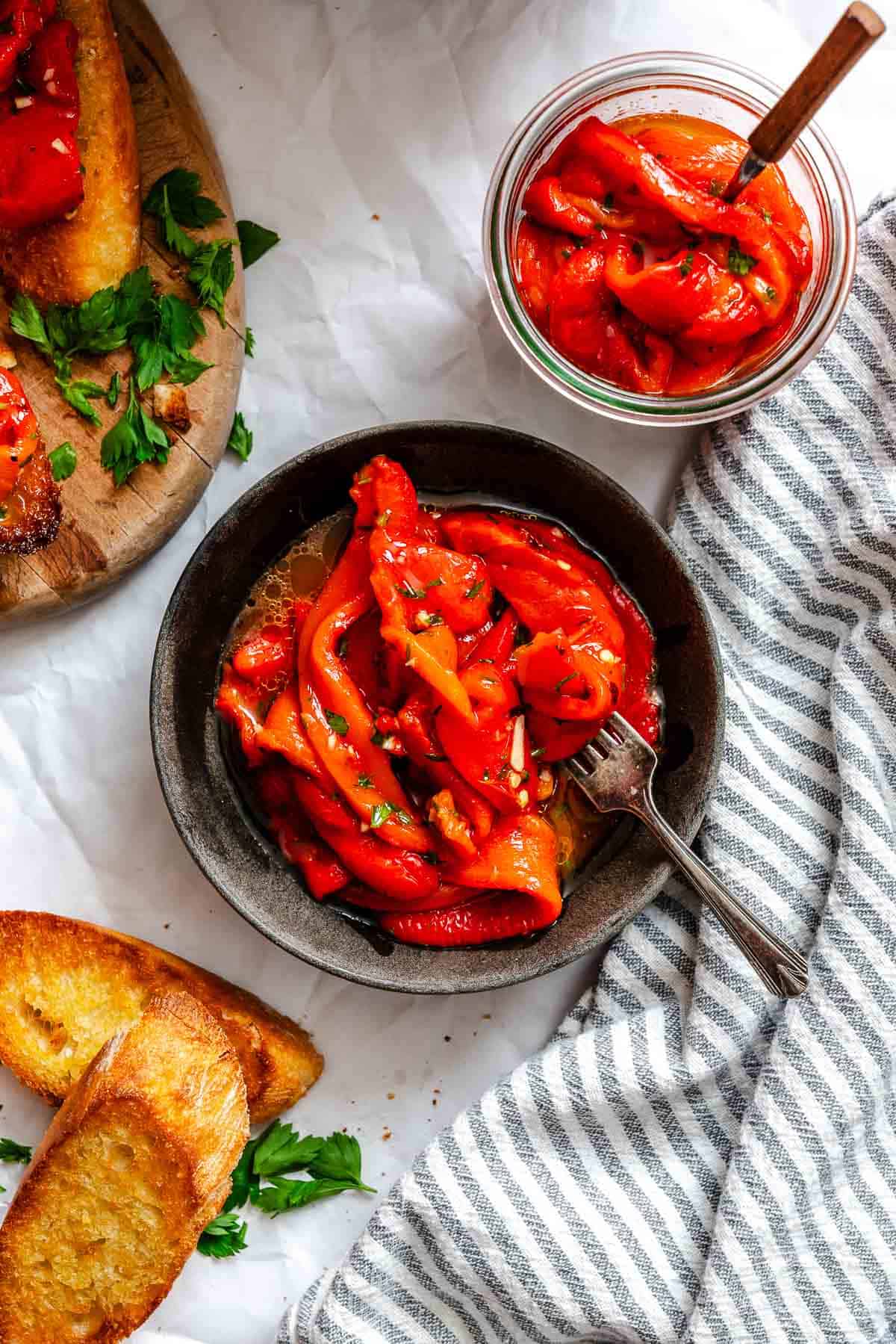 Overhead view of a small black bowl filled with glossy roasted red pepper strips in marinade, surrounded by a glass jar of peppers, toasted bread, and a striped kitchen towel.
