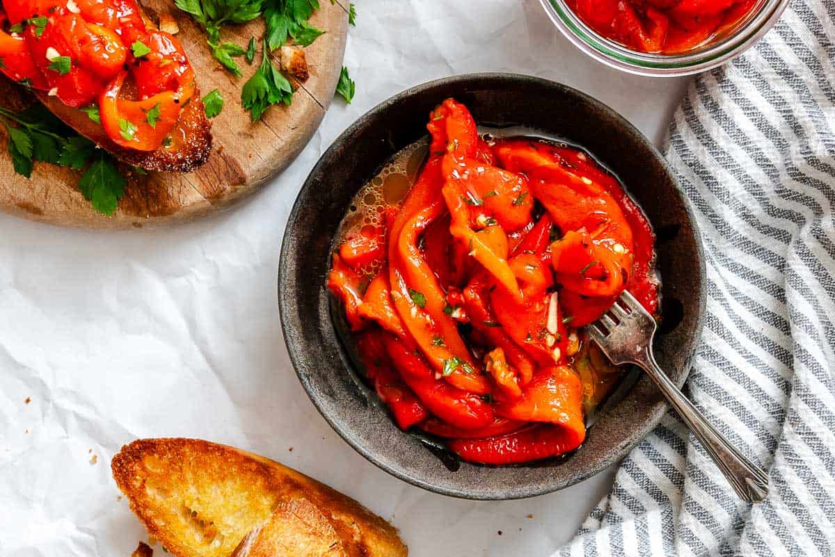 Horizontal layout showing a bowl of marinated roasted red peppers with a fork, partially visible crostini on the side, and a rustic kitchen towel nearby.