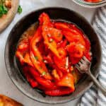 Close-up shot of a dark ceramic bowl filled with roasted red pepper strips marinated with olive oil, garlic, and herbs, with a fork resting inside the bowl.
