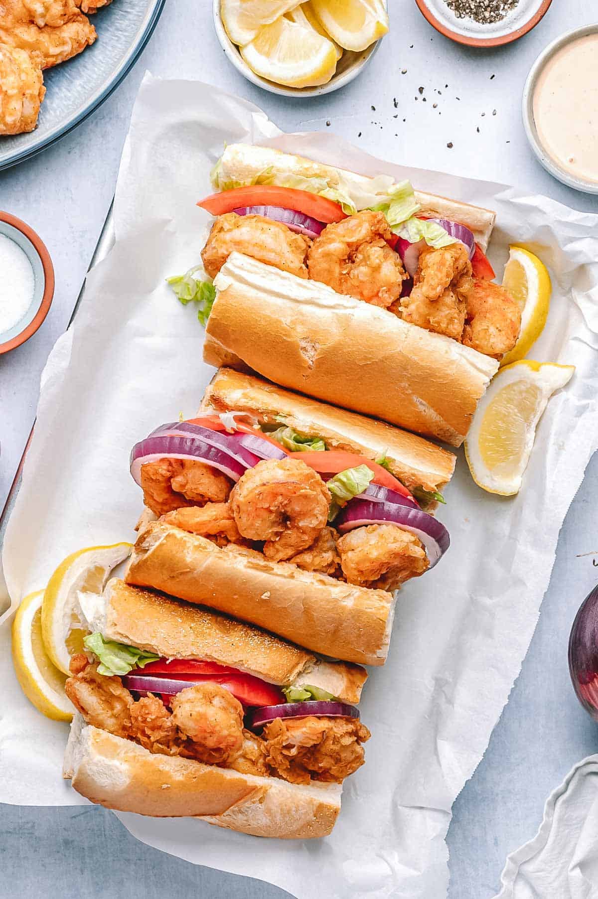Overhead view of three shrimp po'boy sandwiches stuffed with crispy shrimp, sliced tomato, lettuce, and red onion on toasted French bread, garnished with lemon wedges and served with sauce.