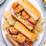 Shrimp po'boys served on a white plate with crispy potato chips. Sandwiches are filled with fried shrimp, tomatoes, lettuce, and red onion, with lemon wedges and creamy sauce on the side.