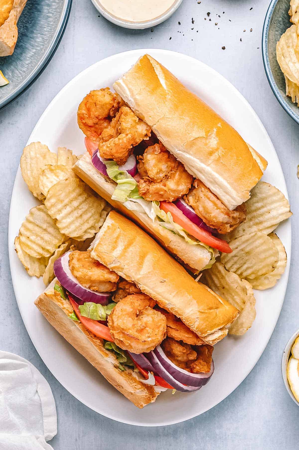Shrimp po'boys served on a white plate with crispy potato chips. Sandwiches are filled with fried shrimp, tomatoes, lettuce, and red onion, with lemon wedges and creamy sauce on the side.