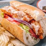 Close-up of a single shrimp po'boy sandwich on a plate with chips. The sandwich is drizzled with creamy sauce and packed with crispy shrimp, fresh tomato, lettuce, and red onion.