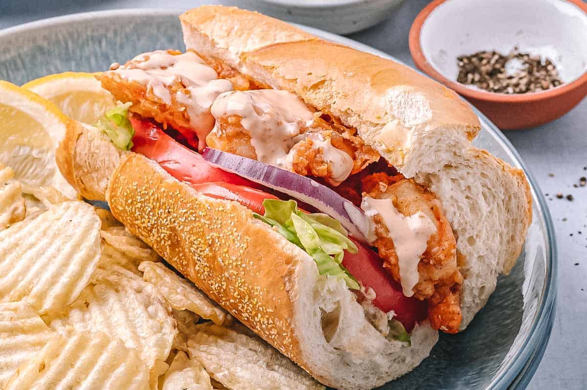 Close-up of a single shrimp po'boy sandwich on a plate with chips. The sandwich is drizzled with creamy sauce and packed with crispy shrimp, fresh tomato, lettuce, and red onion.